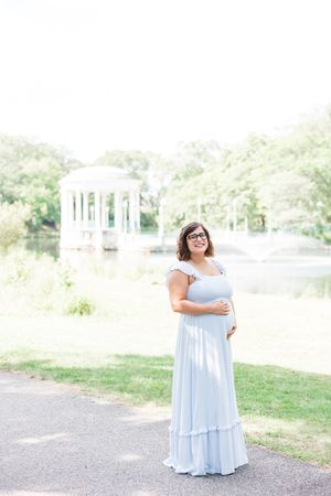 Person in a flowing light blue dress poses gracefully outdoors near a serene lake surrounded by trees and greenery.