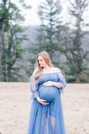 A pregnant woman in a flowing blue off-shoulder dress poses in a natural outdoor setting among trees.