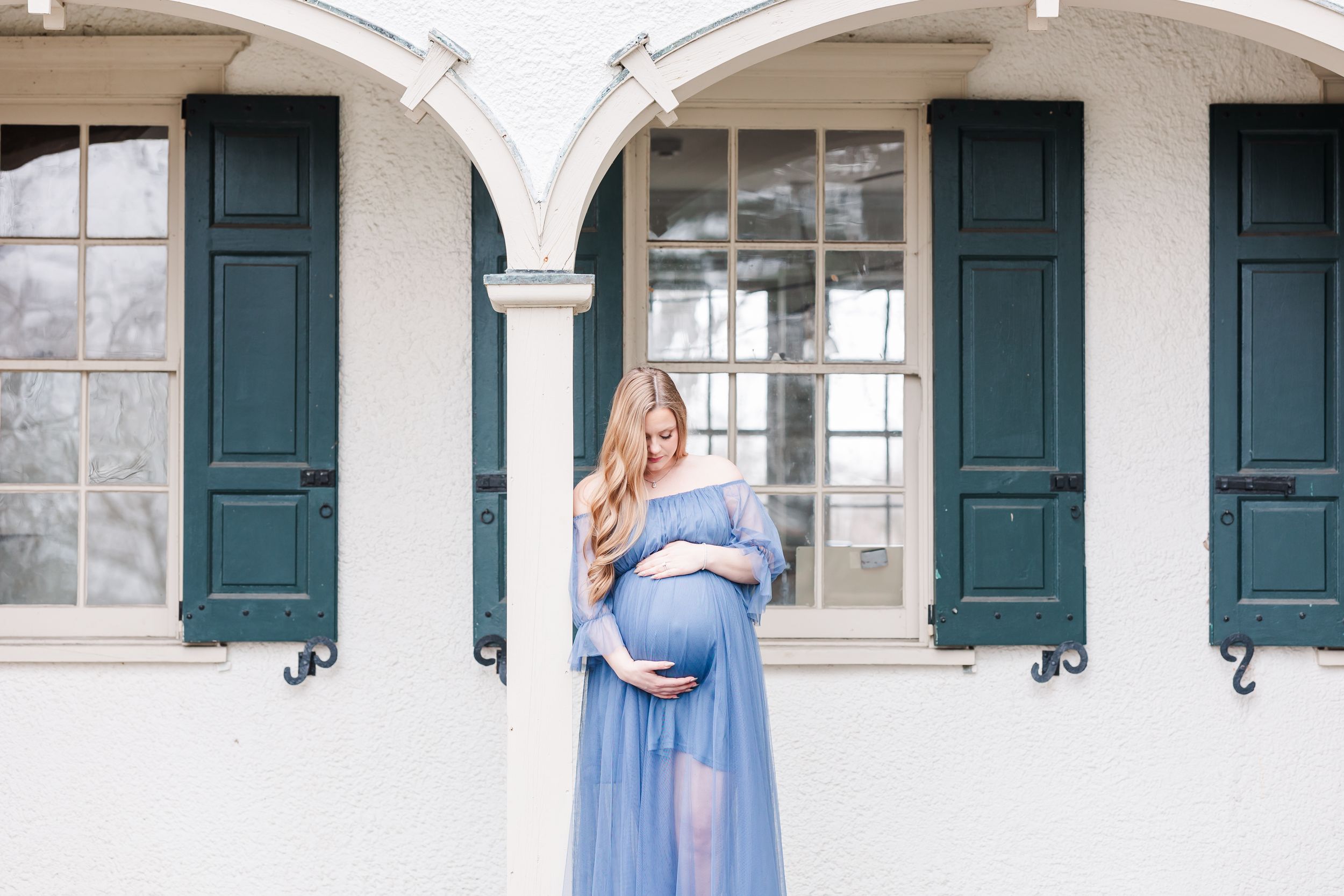 A maternity photo shoot in front of a white arched building with green shutters shows a flowing blue dress.
