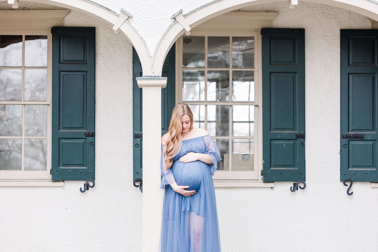A maternity photo shoot in front of a white arched building with green shutters shows a flowing blue dress.