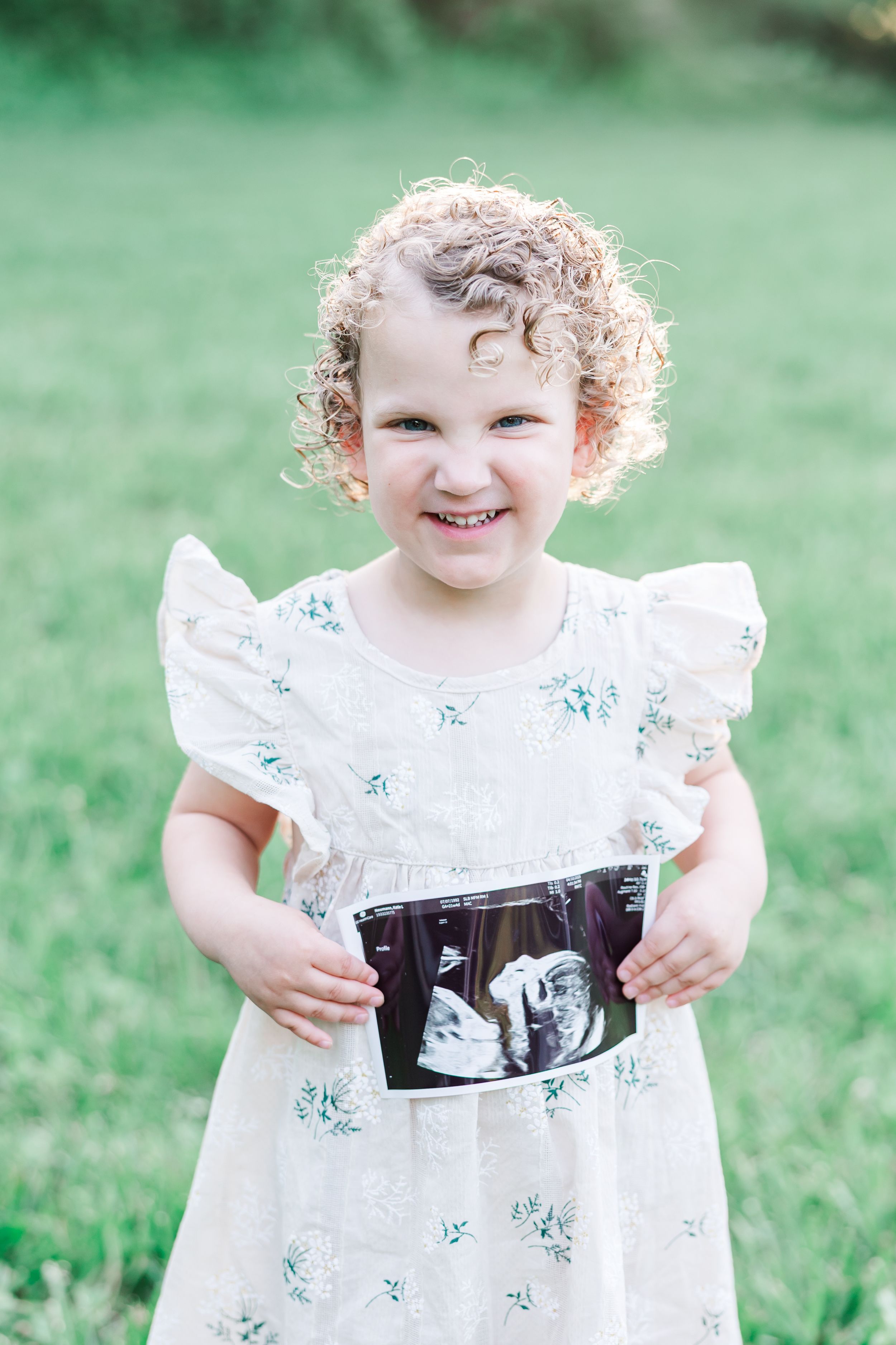 A smiling toddler in a white floral dress holds up an ultrasound photo while standing on grass.
