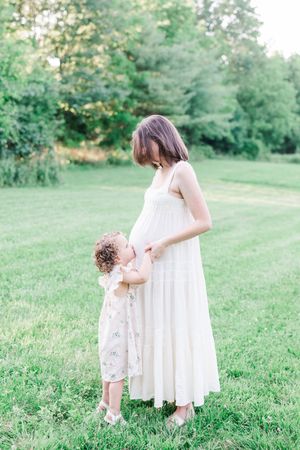 Two figures in soft white dresses embrace in a gentle moment on a sunlit grassy hillside.