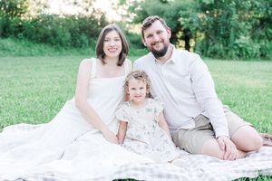 A family in white outfits sits together on a blanket in a lush green field during a sunny outdoor photo session.