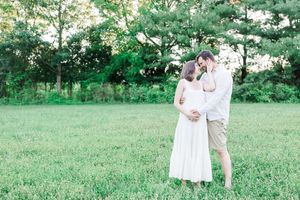Couple dressed in white embracing in a lush green meadow surrounded by trees in a romantic outdoor setting.