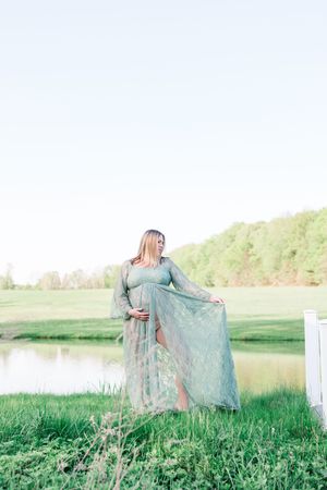 Woman in flowing teal dress walks through serene spring meadow at golden hour near lake.