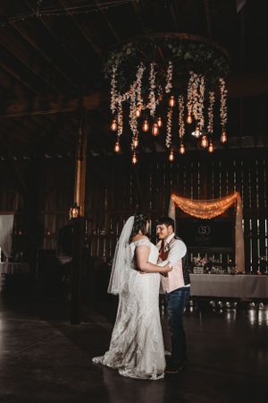 Couple dancing in a rustic barn venue decorated with hanging lights and floral arrangements.