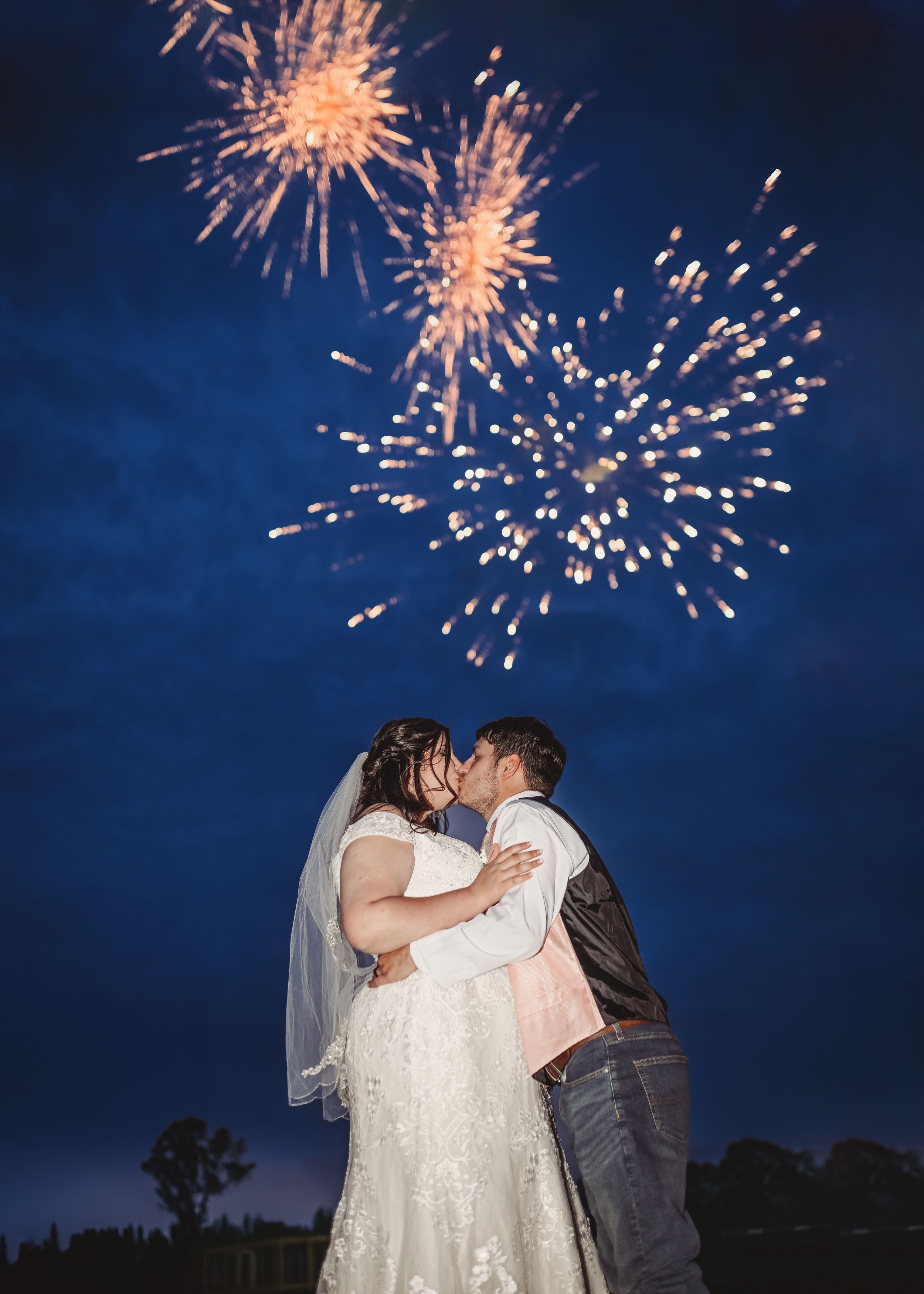 Newlyweds share a kiss under spectacular fireworks display in night sky.