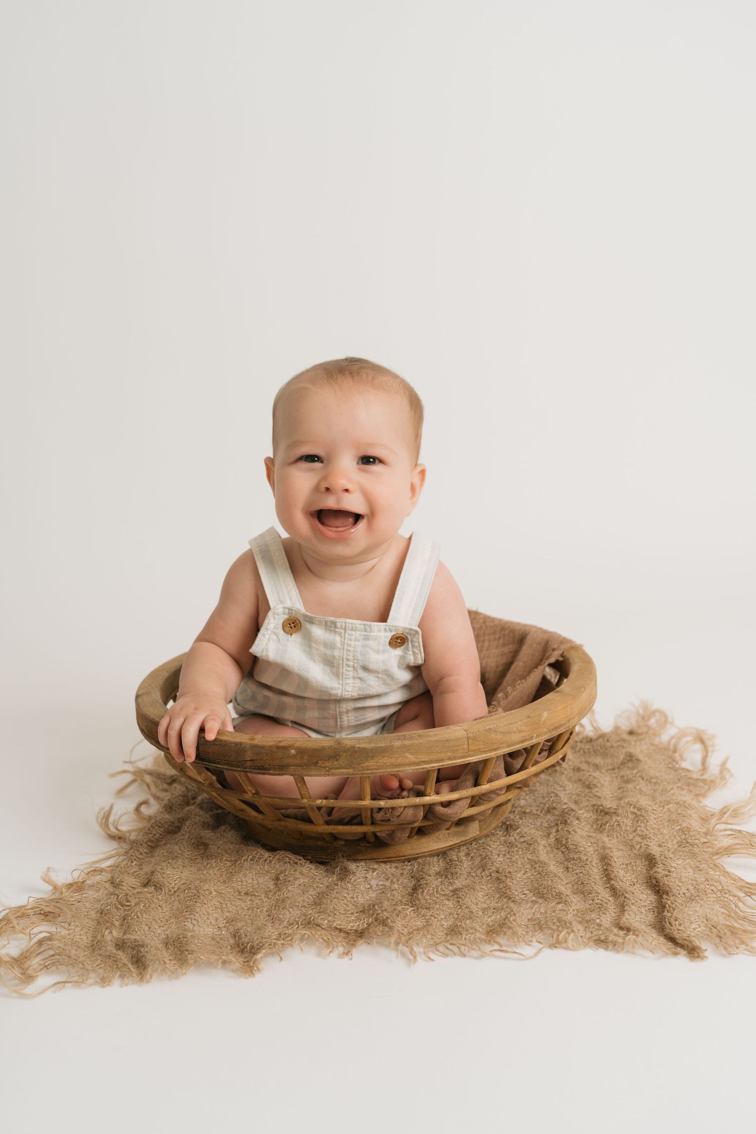 A small baby sits in a wooden bowl on burlap fabric, showing a progression of joyful expressions.