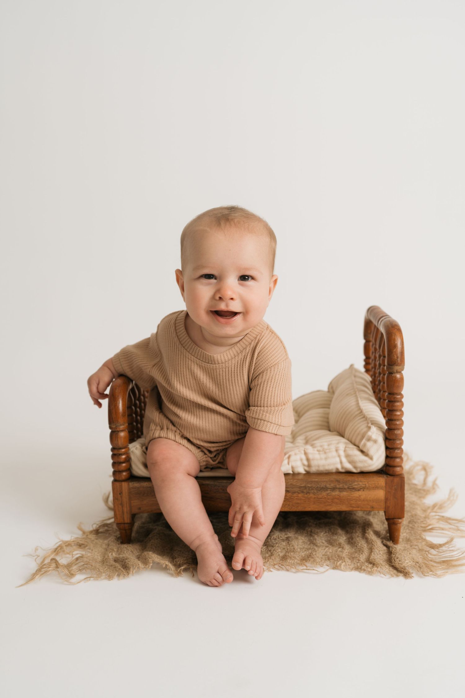 A baby in a tan ribbed outfit sits on a wooden doll bed with white bedding in a minimalist studio setting.