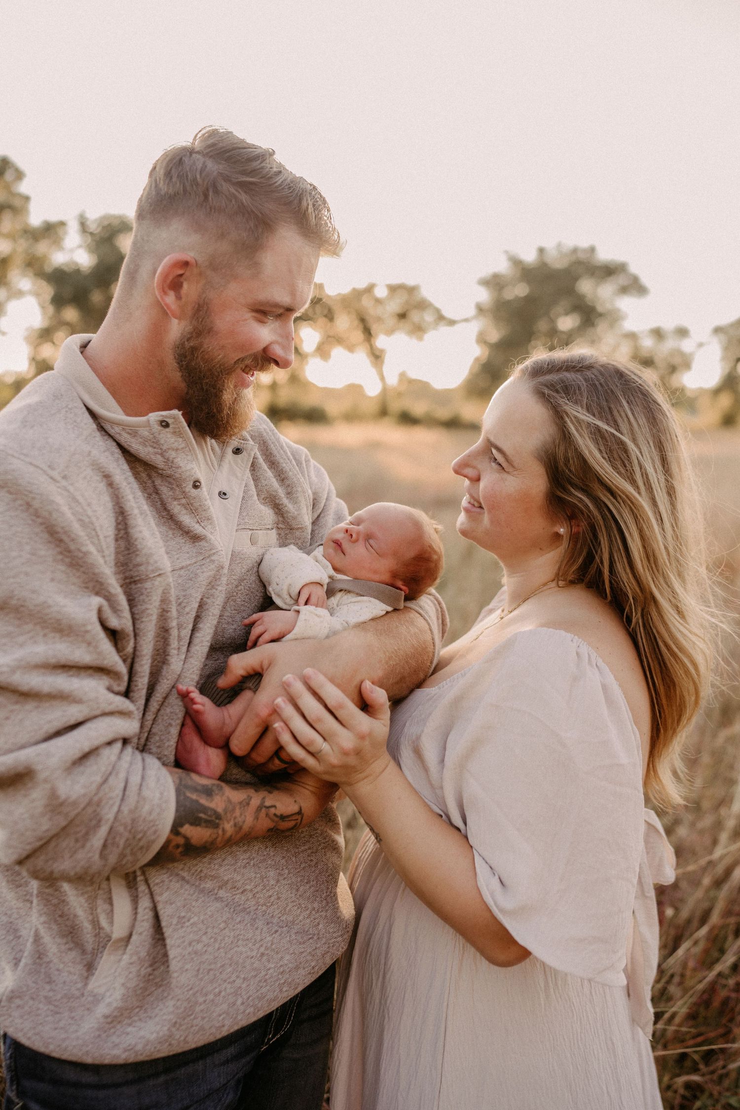A couple shares a tender moment with their newborn baby outdoors during golden hour in a natural field setting.