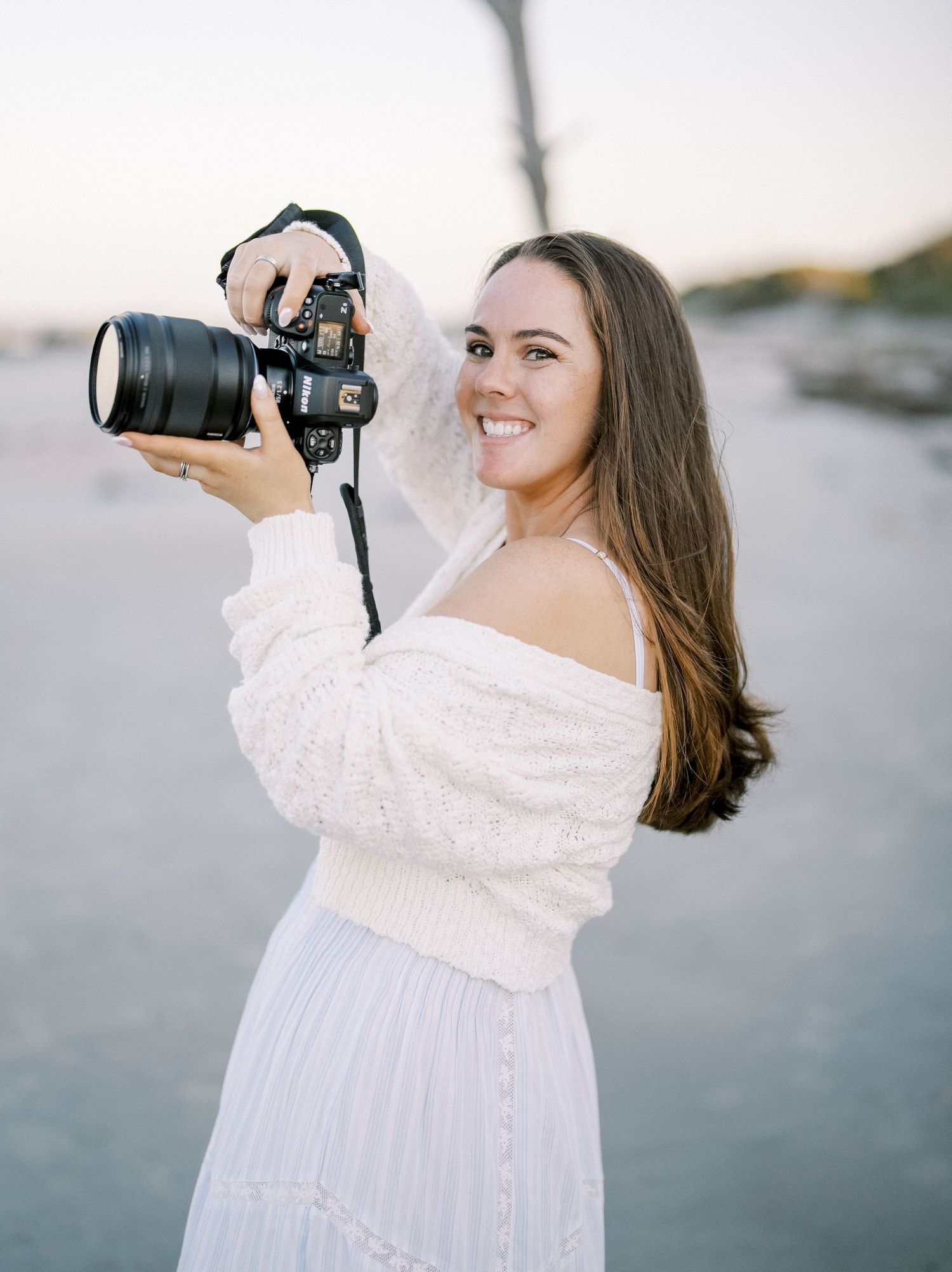 Photographer in white dress holding professional camera outdoors during golden hour.