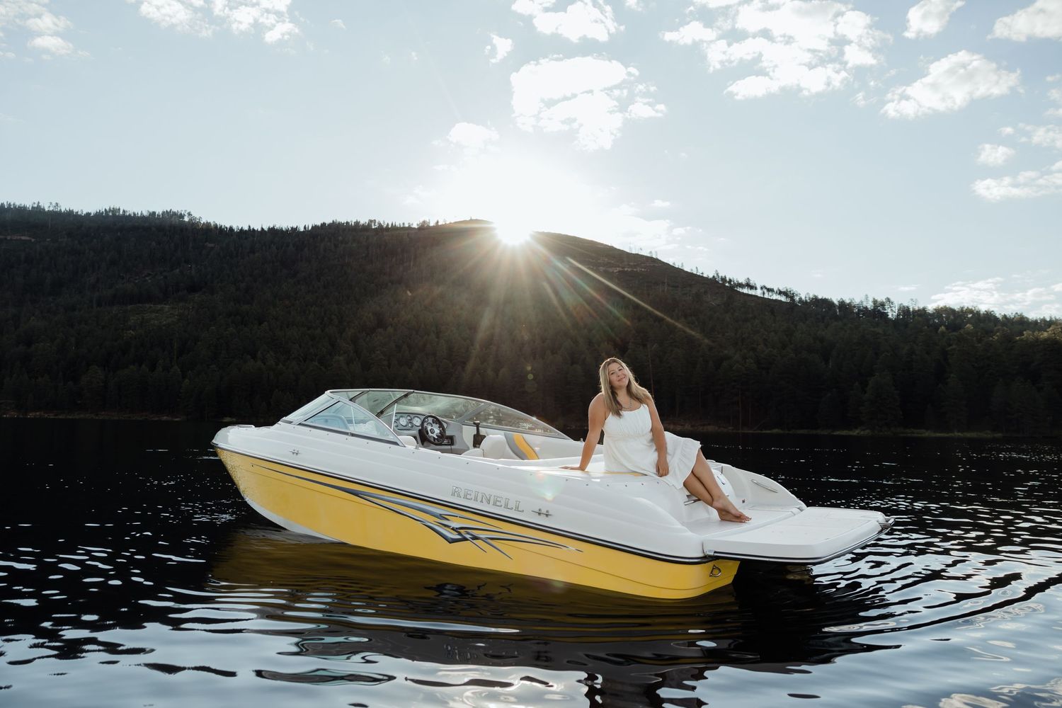Woman in white dress sitting on yellow speedboat on calm lake with mountain backdrop at sunset.