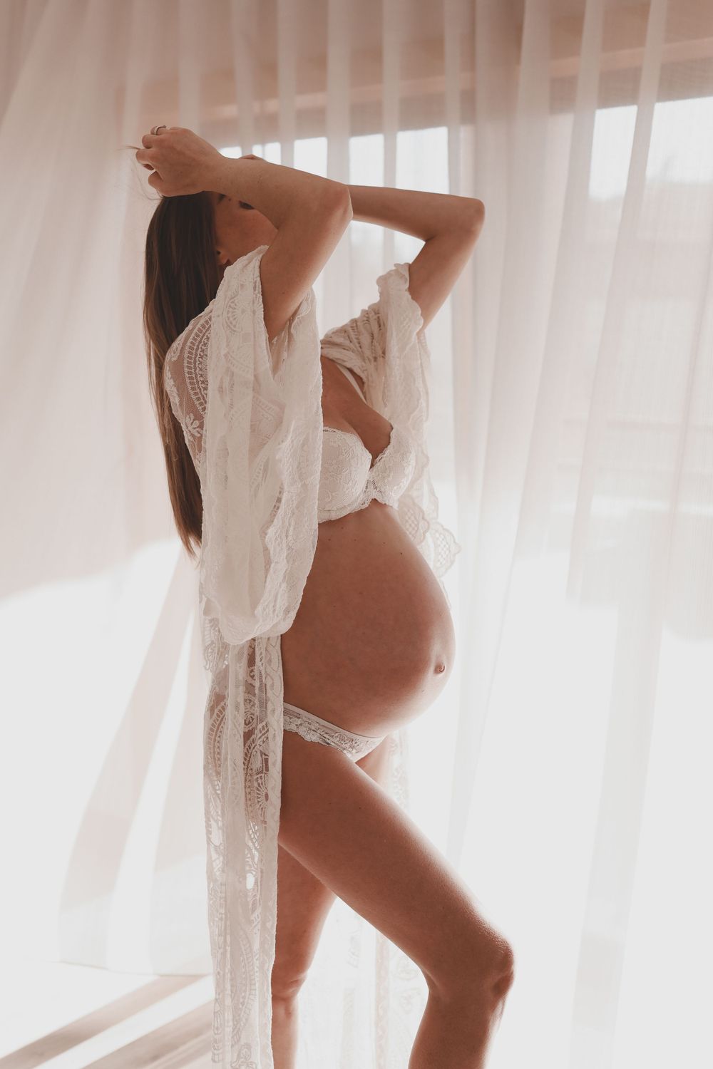 Pregnant woman in white lace robe and bra posing by sheer curtains in soft natural light.