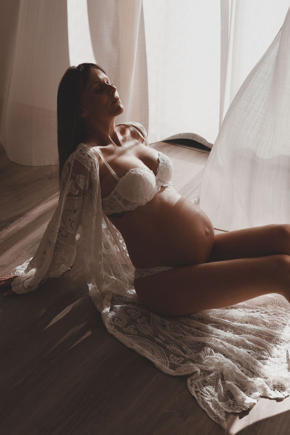 Pregnant woman in white lace robe sitting on floor by window in warm natural light.