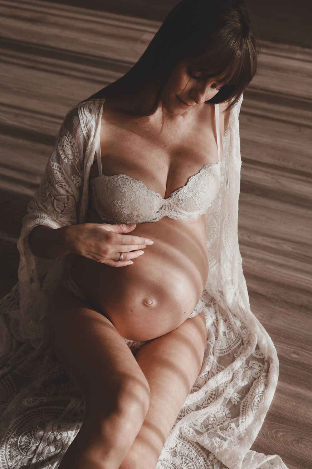 Close-up of pregnant woman in white lace robe cradling baby bump in warm light.