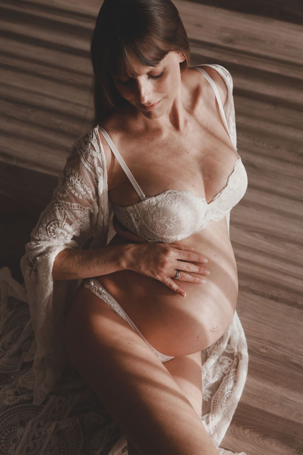Pregnant woman in white lace lingerie sitting and gently holding baby bump indoors.