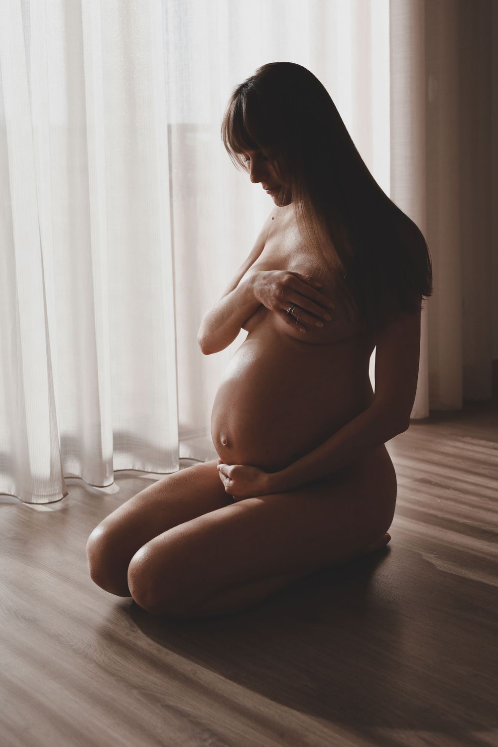 Pregnant woman kneeling nude on wooden floor by sheer curtains in soft natural light.
