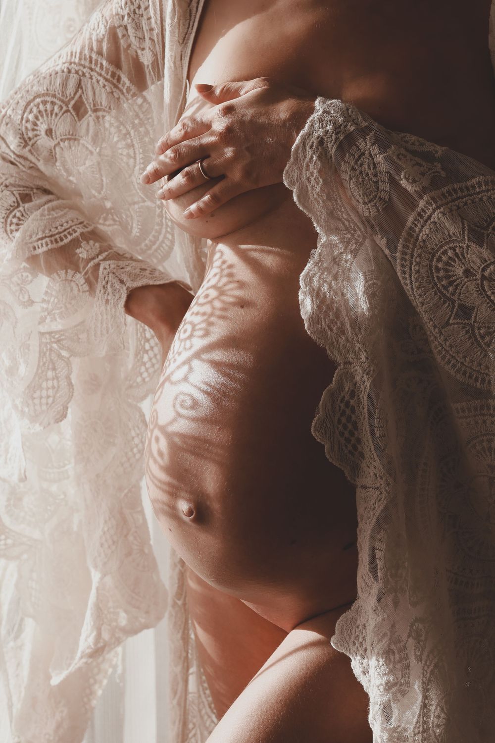 Close-up of pregnant belly with lace robe open, showing intricate shadow patterns on skin.