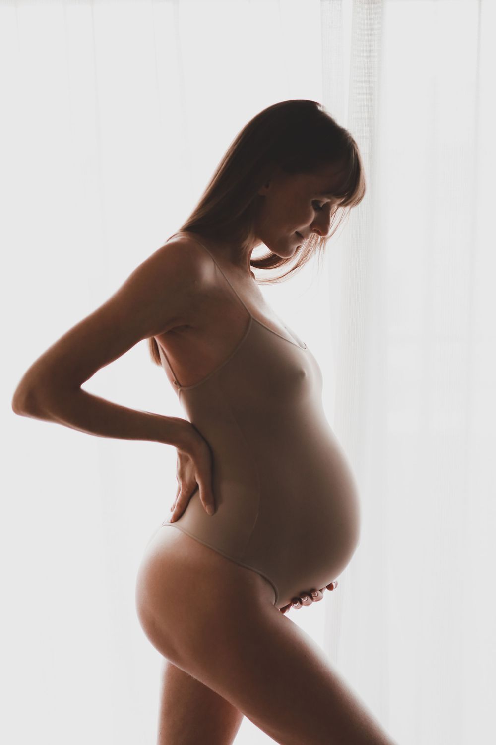 Silhouette of pregnant woman in nude bodysuit posing by bright white curtained window.