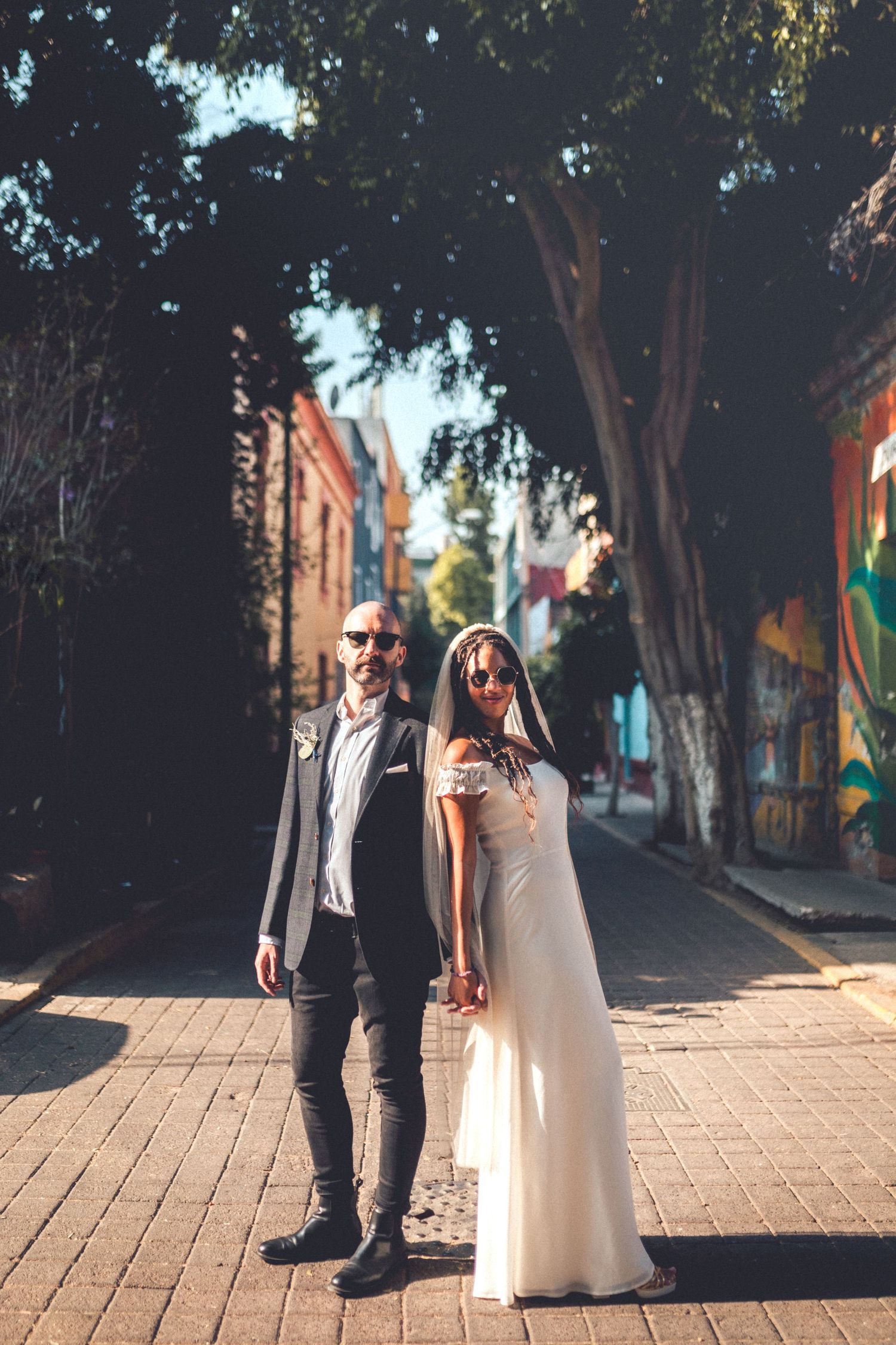 Stylish wedding couple walking down a tree-lined urban street with colorful murals.