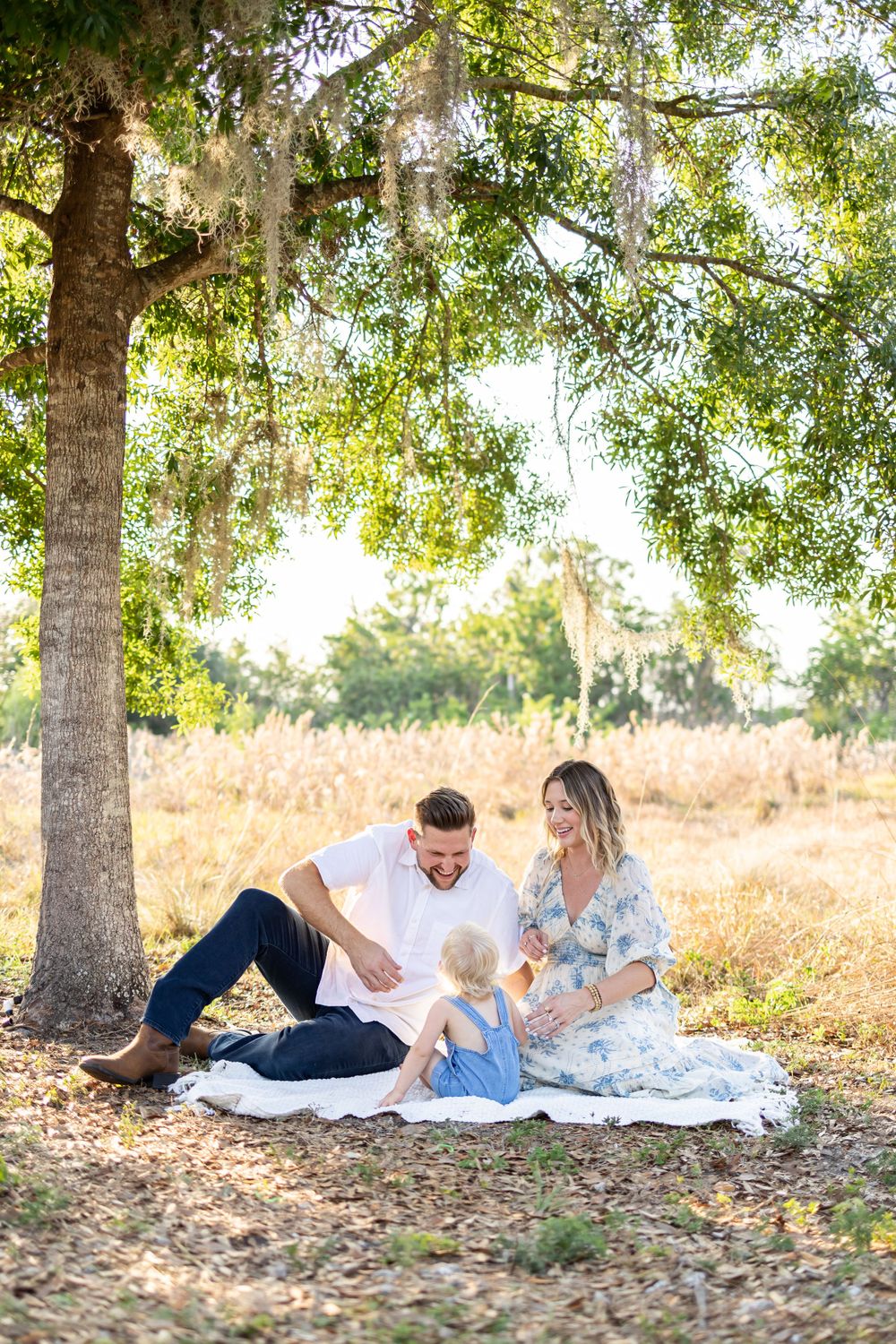 Family enjoys playful moments together on white blanket during outdoor photoshoot.