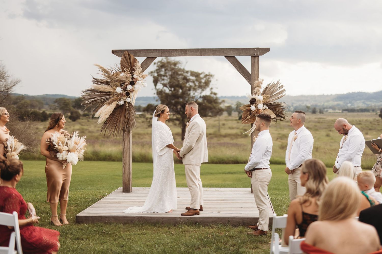 Outdoor wedding ceremony under wooden arch decorated with dried pampas grass arrangements.