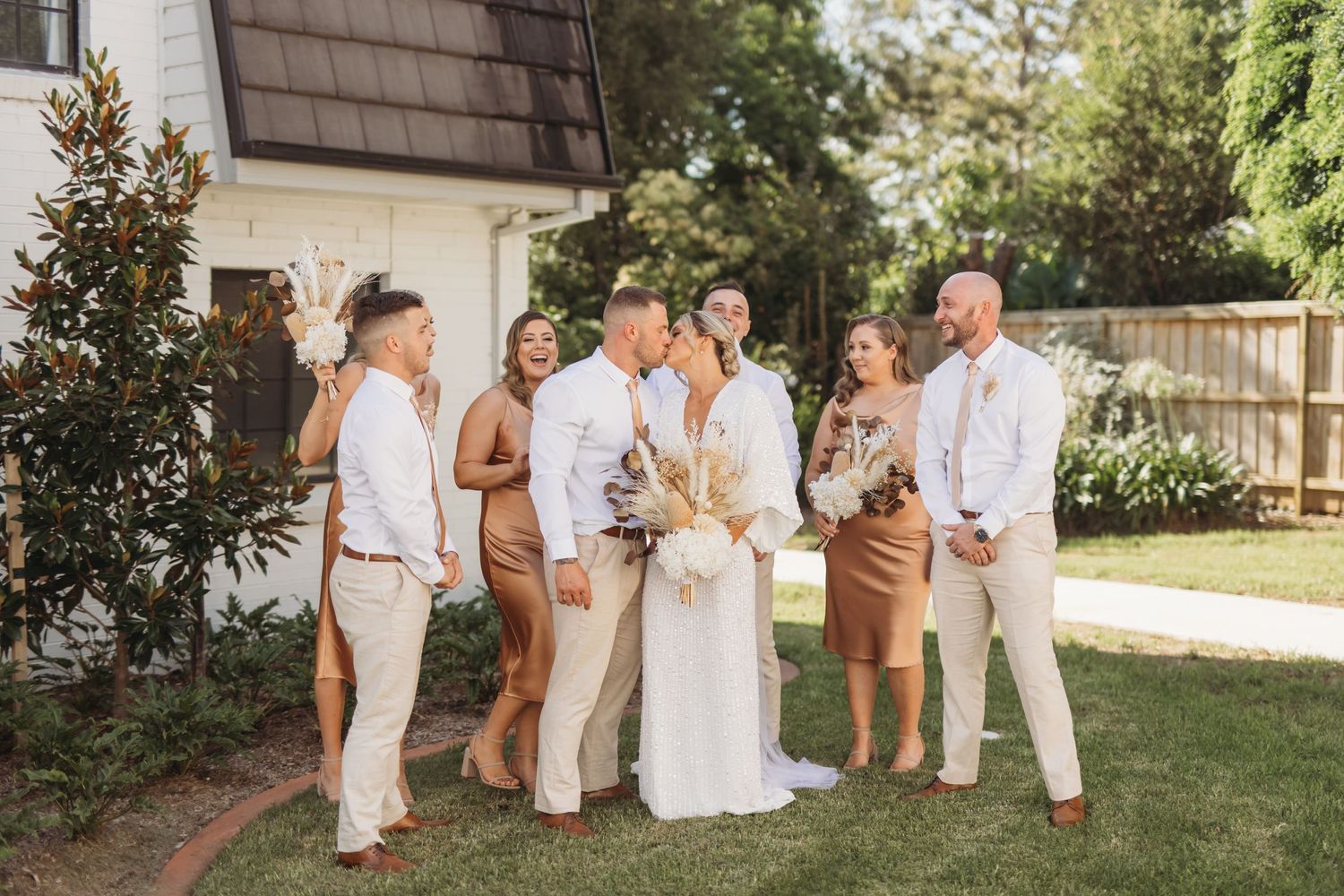 A wedding party poses together in casual attire on a sunny lawn.