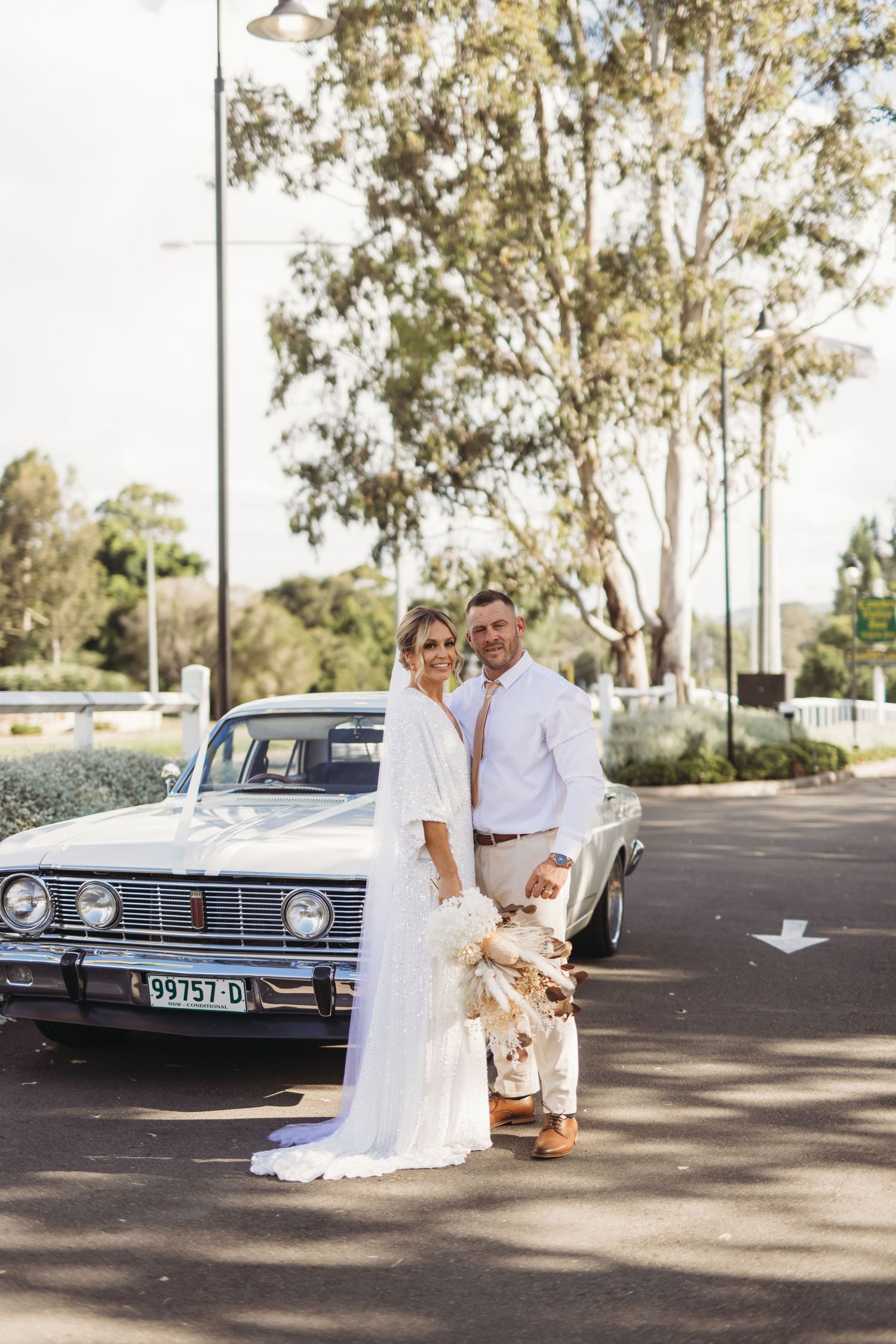 A wedding couple poses with a classic vintage car under tall eucalyptus trees.