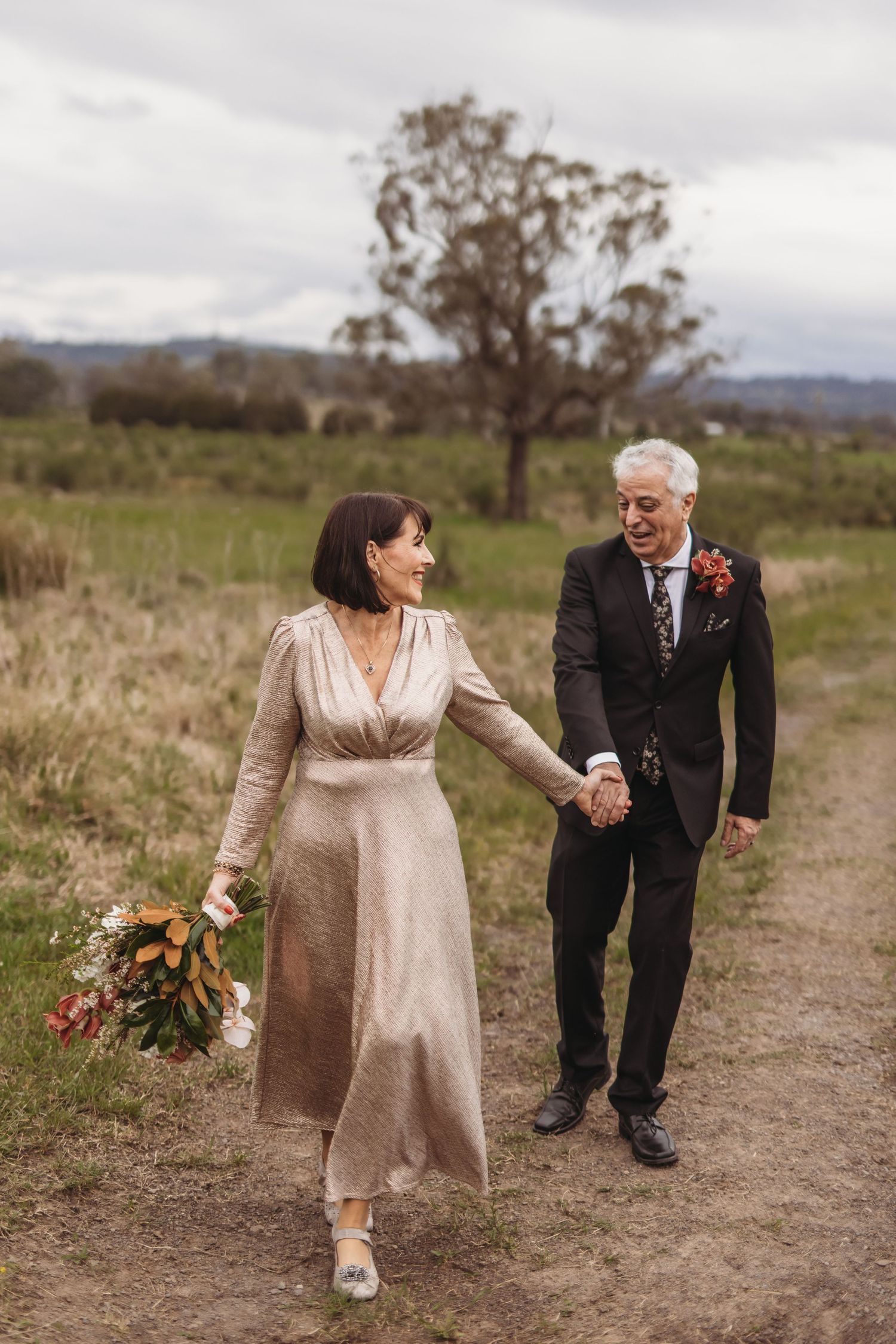 Couple walking hand in hand down countryside path carrying floral bouquet.