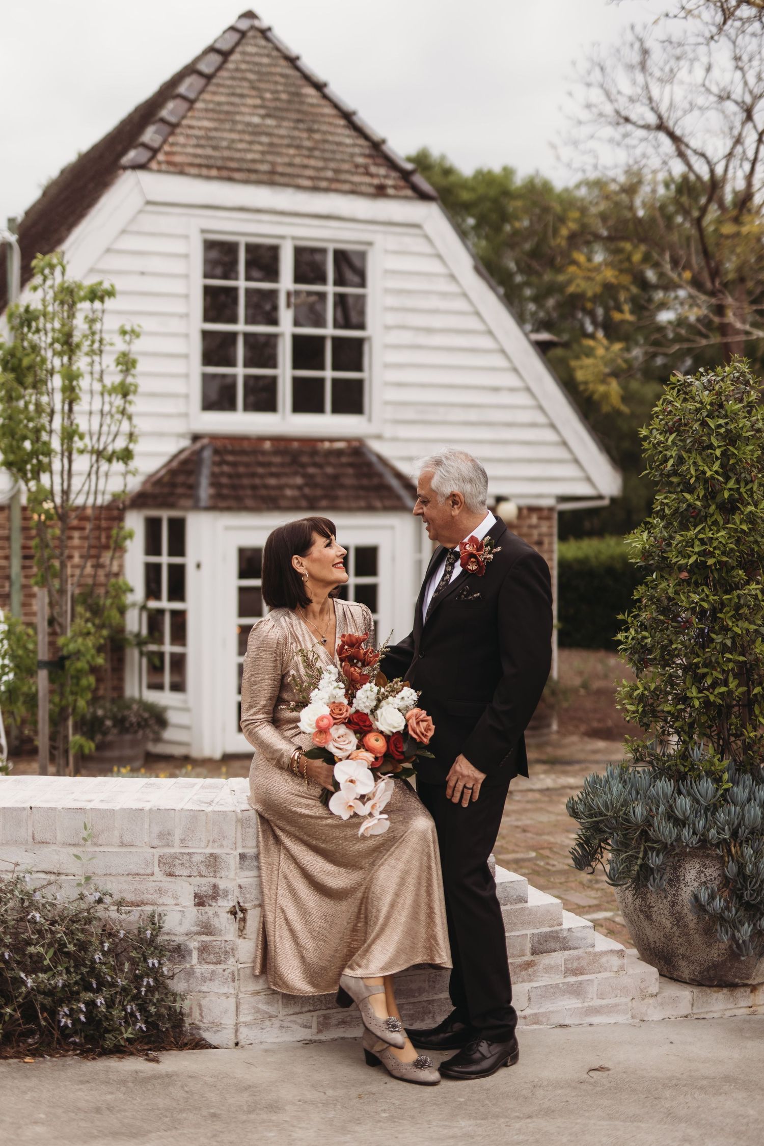 Couple sharing intimate moment in front of charming white cottage with floral bouquet.