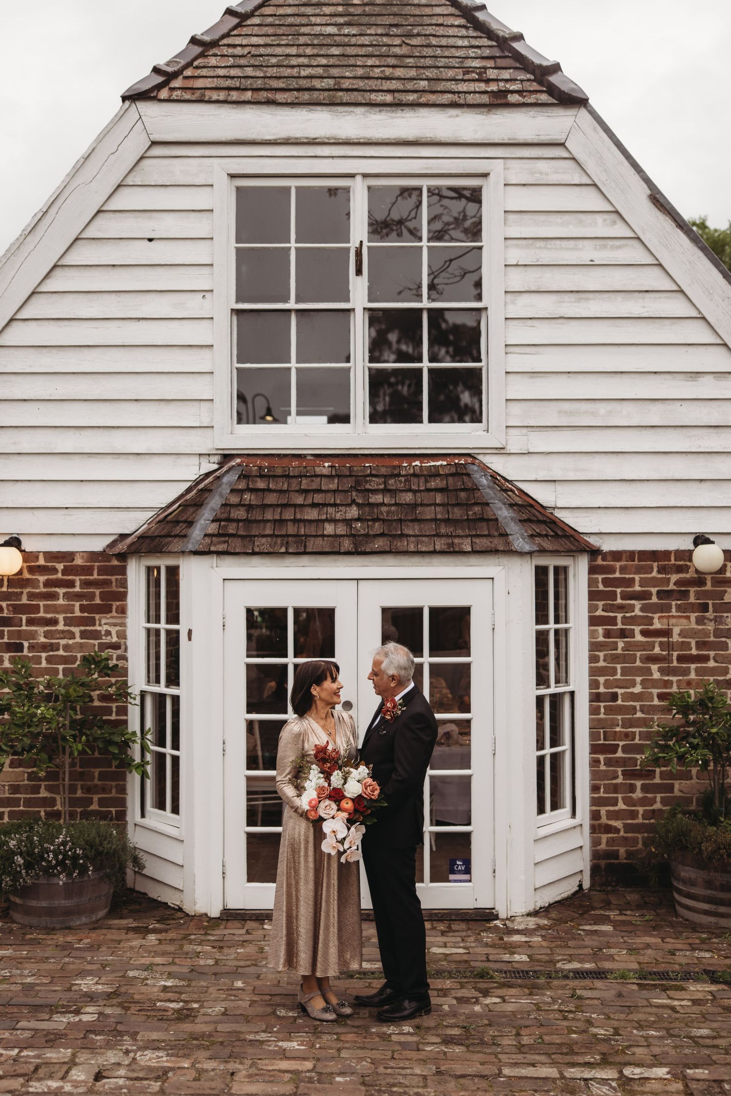 Couple standing in front of white colonial house with bay window and brick accents.