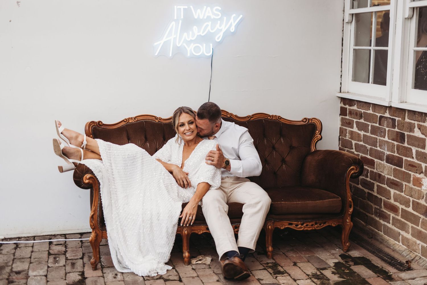 A couple relaxes on a brown vintage couch under neon lighting.