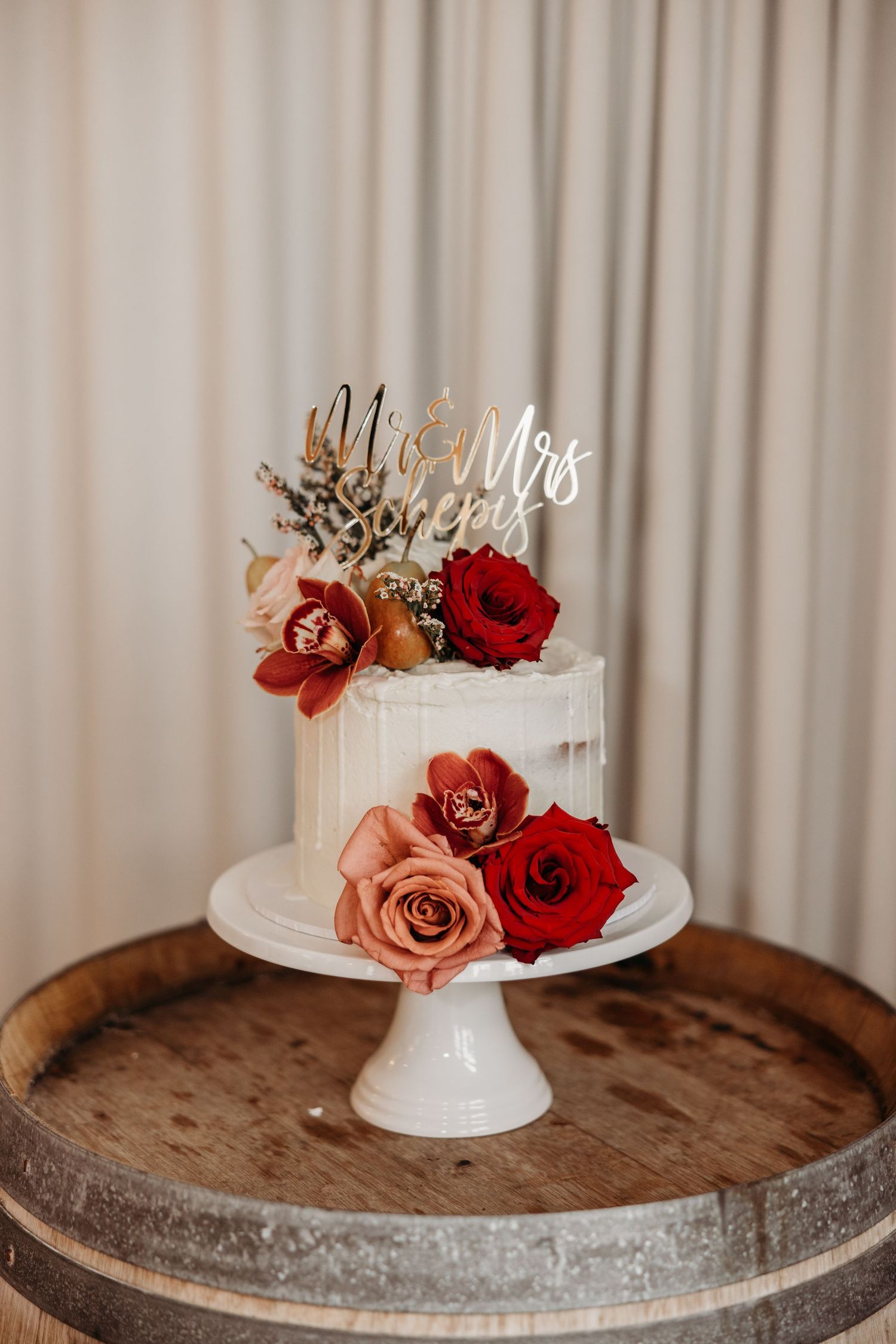White wedding cake with red and pink roses topped with gold Mr and Mrs topper on wooden stand.