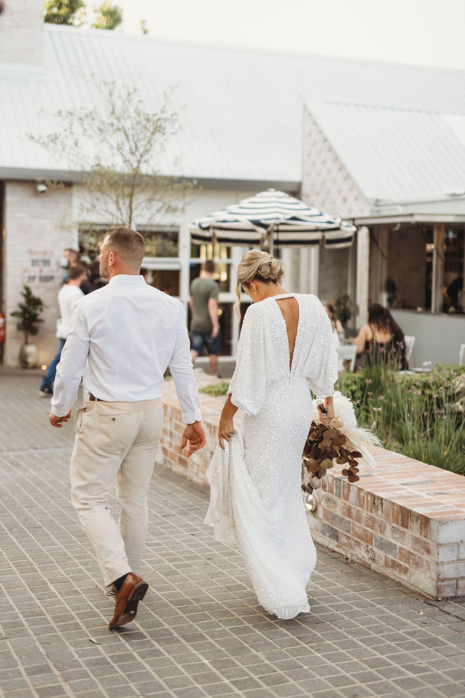 A couple in white wedding attire strolls along a brick pathway near outdoor shops and cafes.