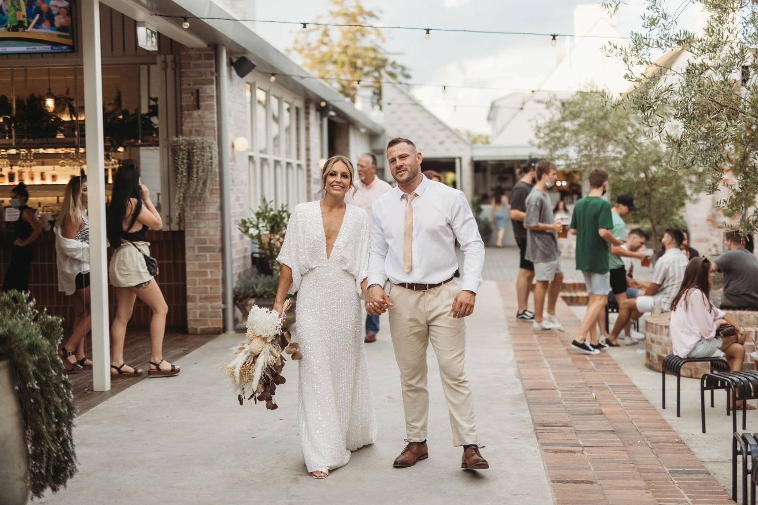 Wedding couple walks hand in hand through a charming outdoor shopping area decorated with string lights.