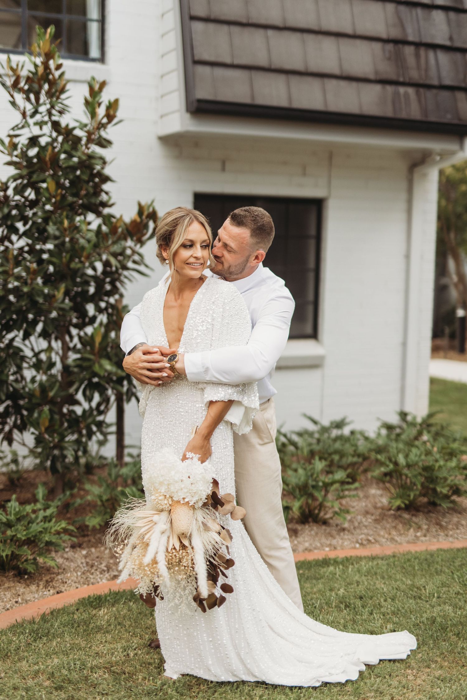 Romantic wedding portrait with dried pampas grass bouquet against white building exterior.
