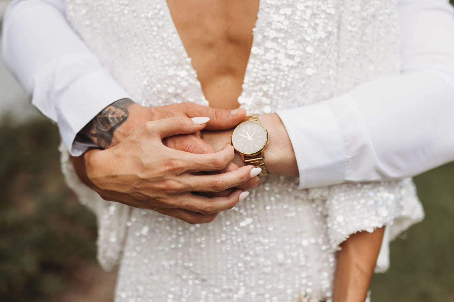 Close-up detail of hands with tattoos and rings against a white lace wedding dress.