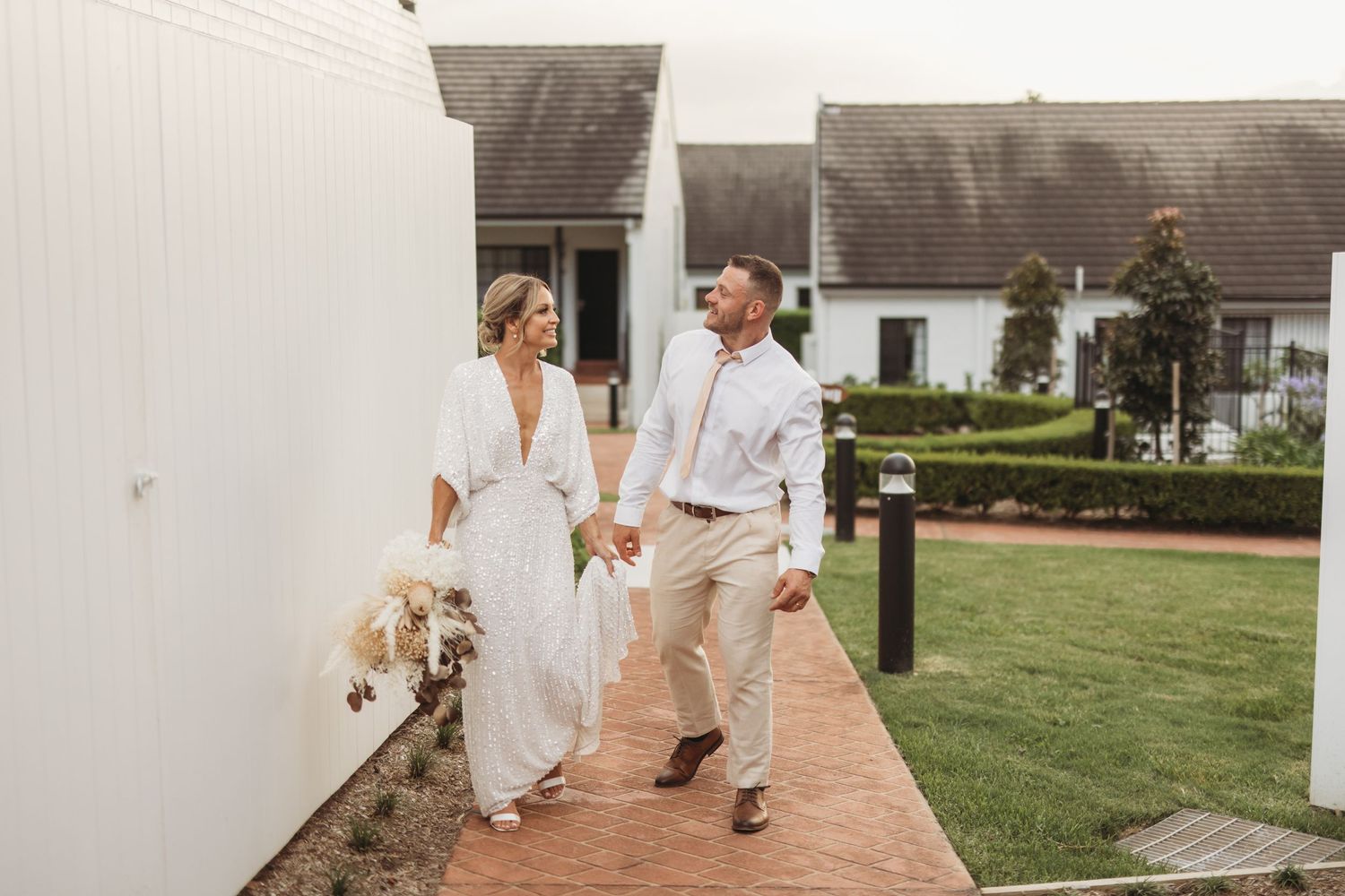 Couple in wedding attire walking along white building path with dried pampas grass bouquet.