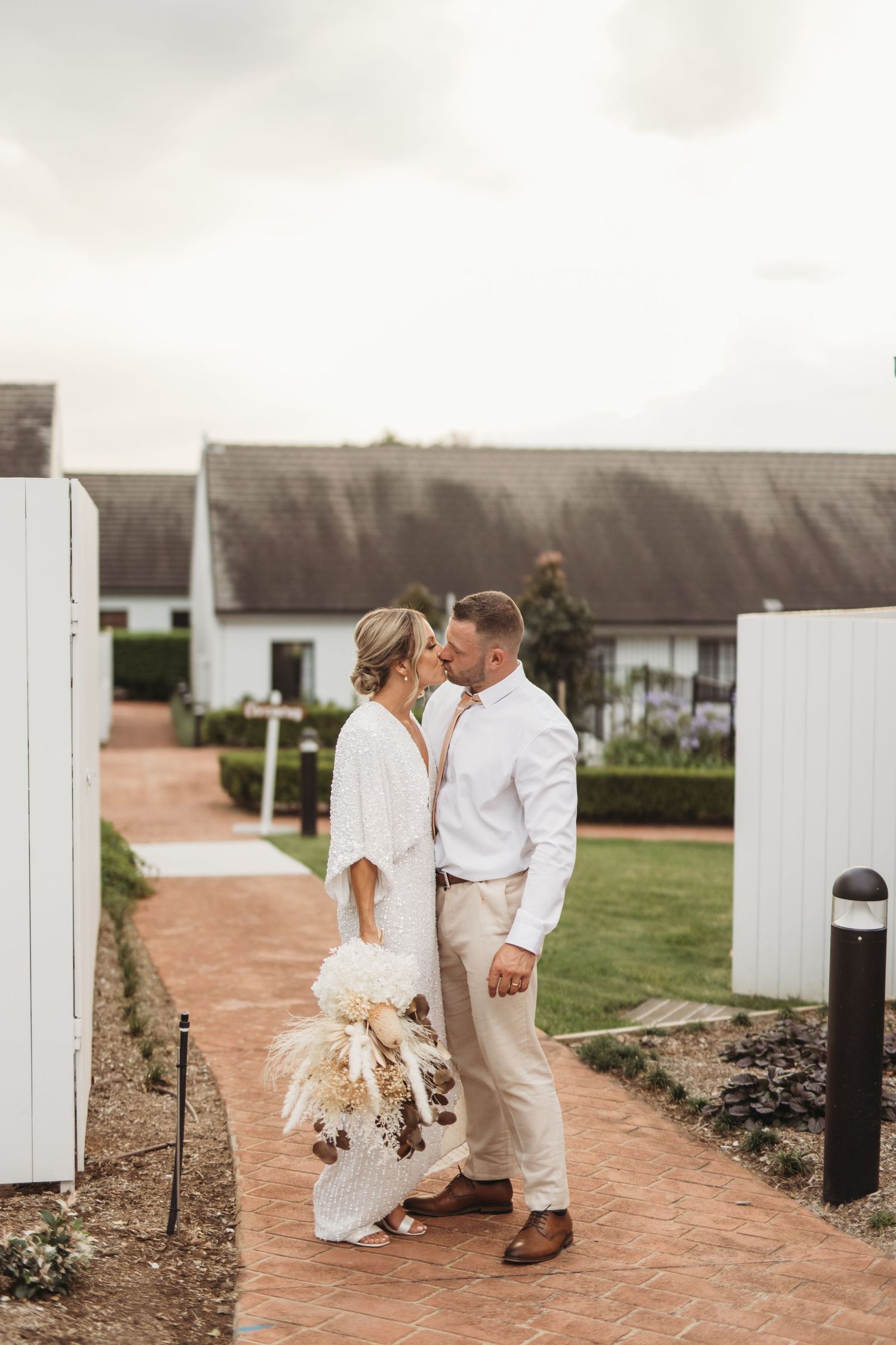 Newlyweds in white and khaki share an intimate moment by a white fence and brick path.