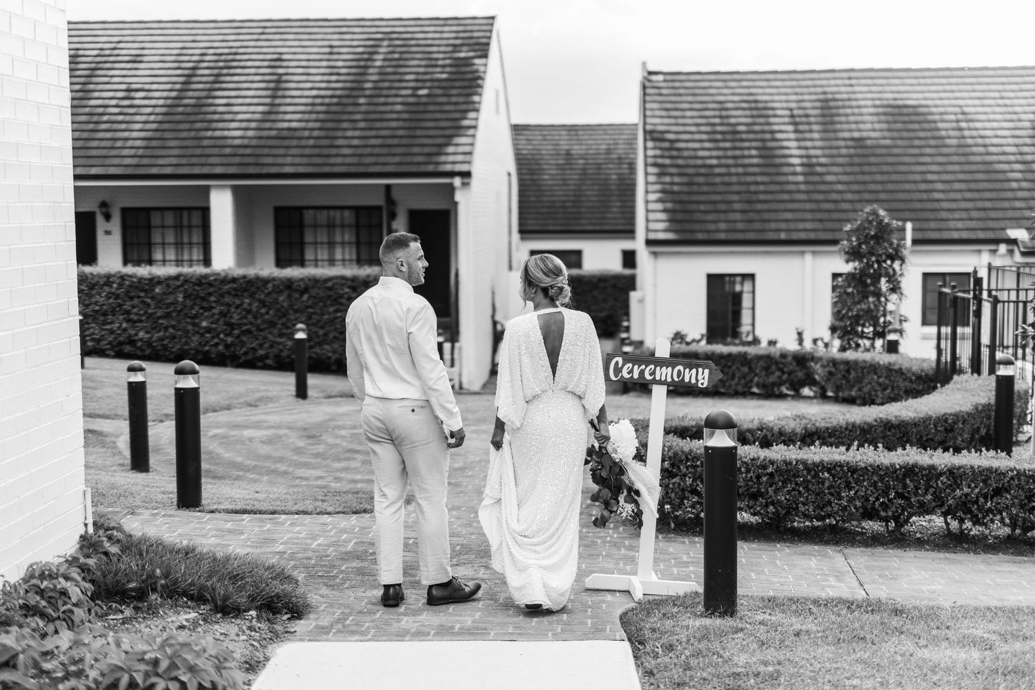A vintage black and white photo showing two figures walking down a path between hedges and houses.
