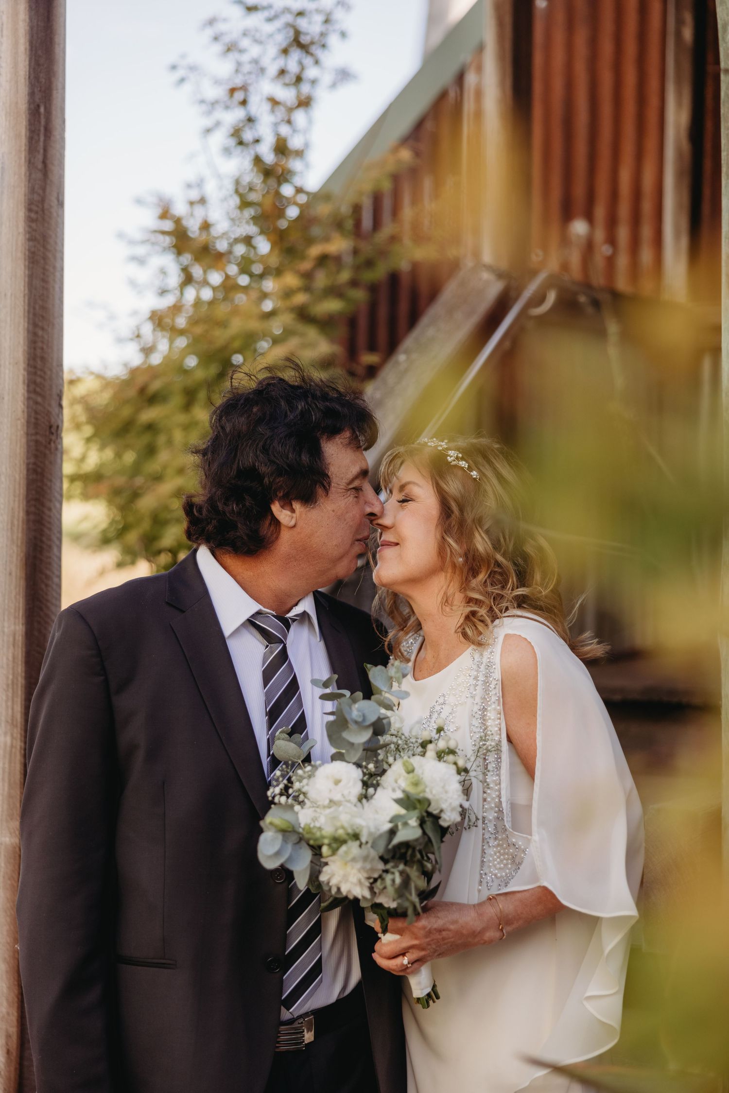 A couple shares an intimate moment while holding a white bouquet against a rustic building exterior.
