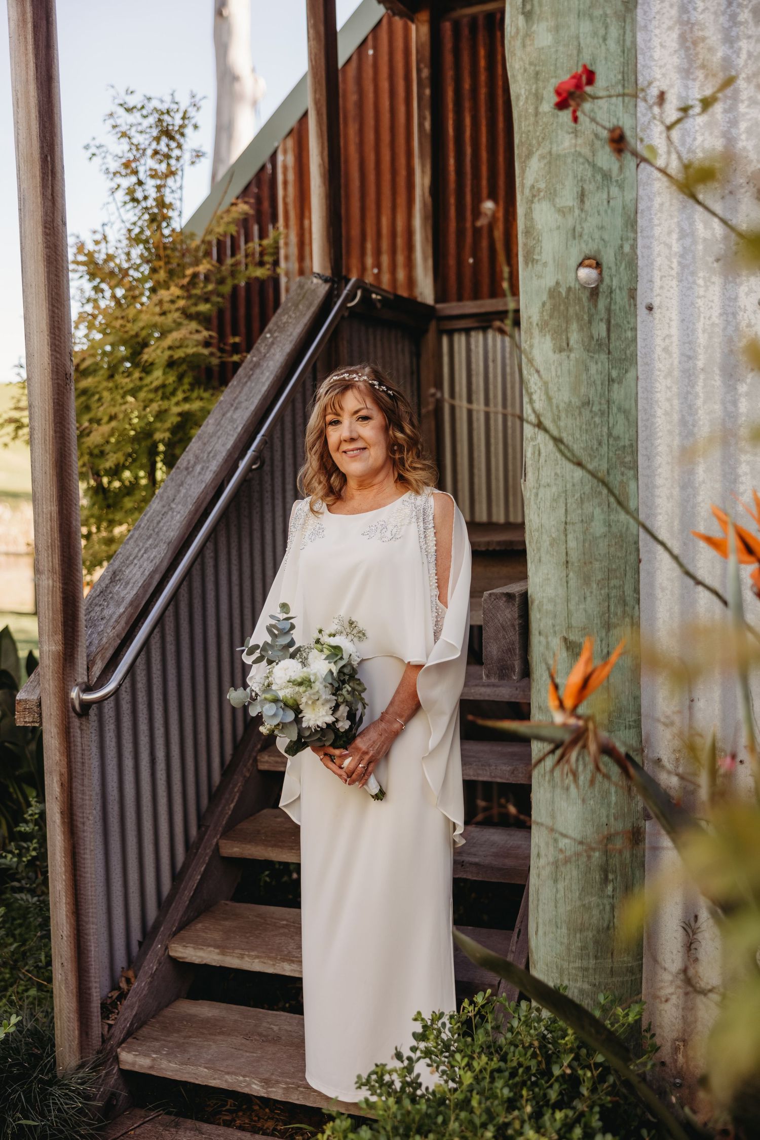A bride in a flowing white dress stands on wooden stairs holding a bouquet of white flowers.