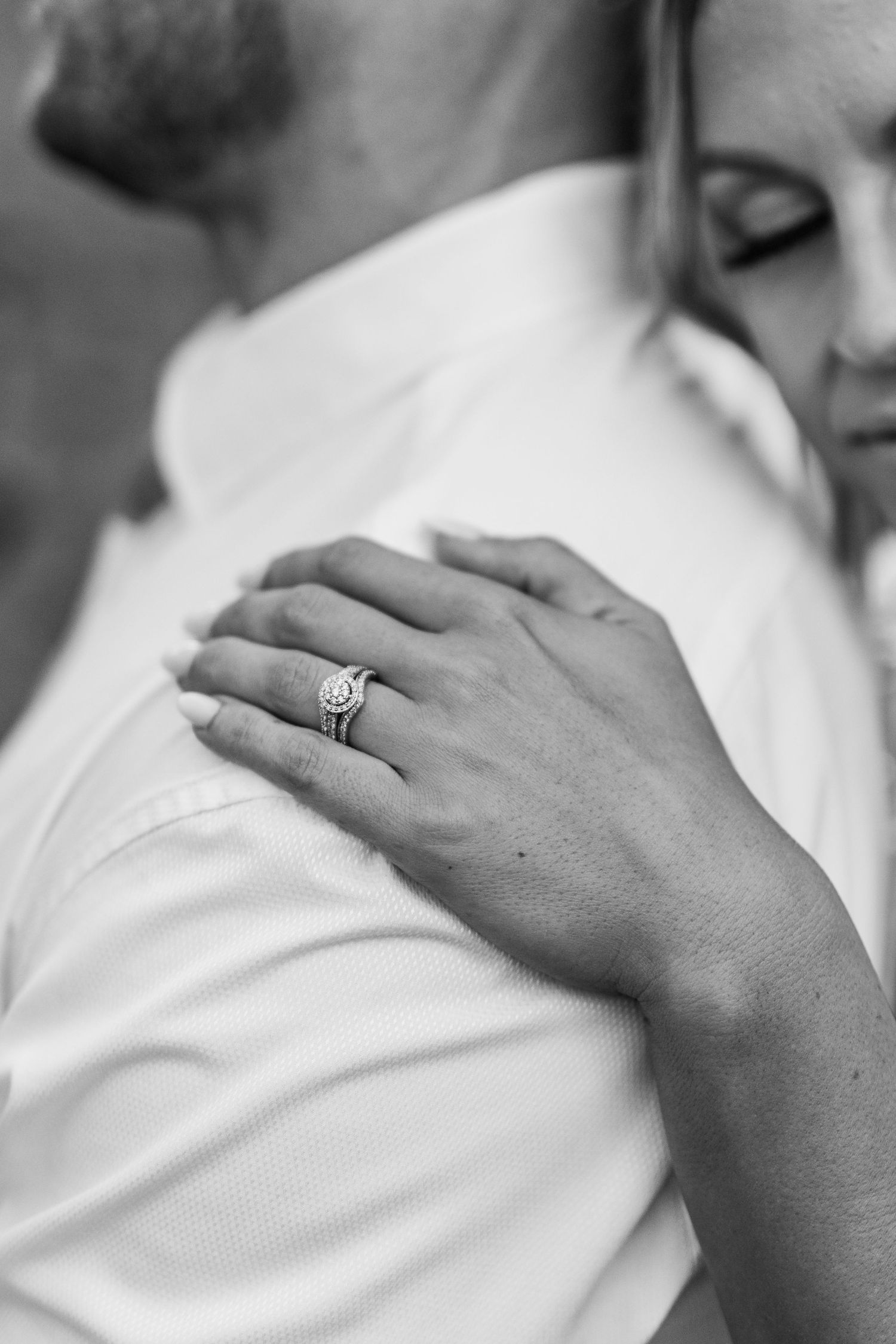 Close-up black and white photo of a hand wearing an engagement ring resting on a white fabric.