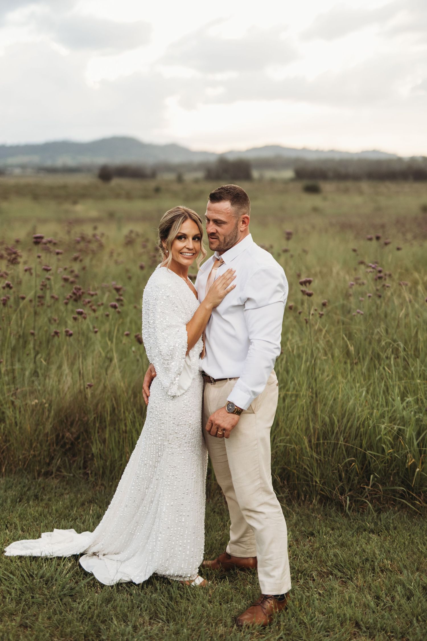 Wedding couple embraces in a rustic field with mountains in the background at sunset.