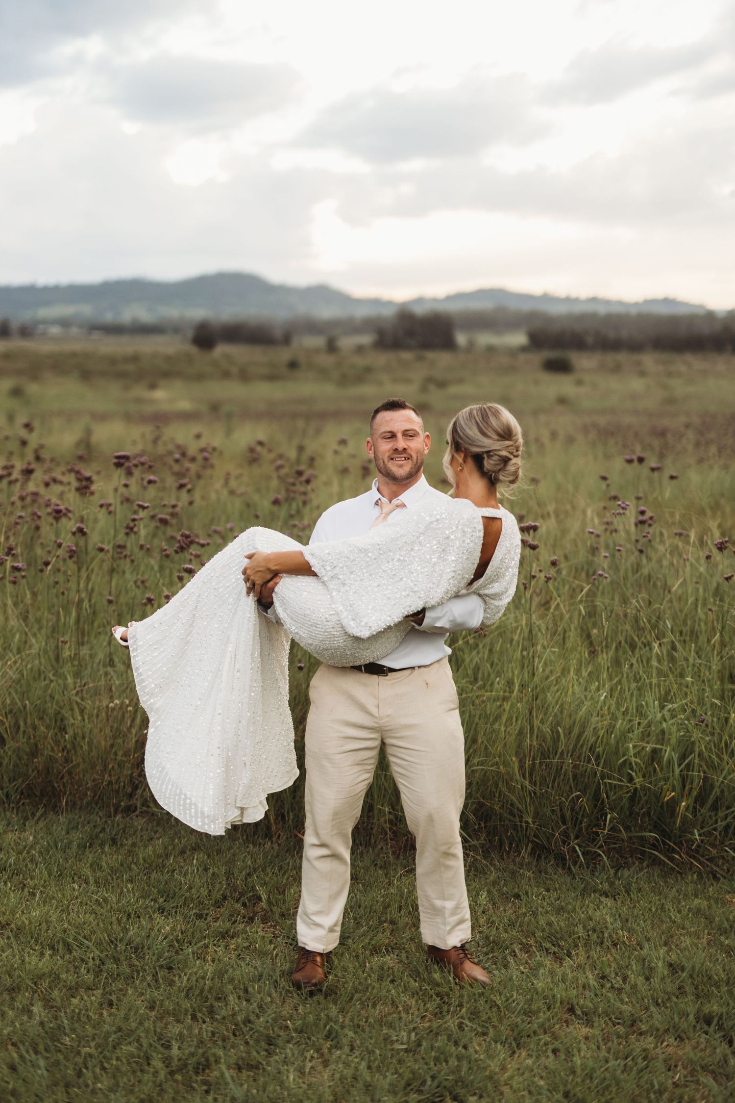 Groom carries bride in her white lace dress against a backdrop of rolling hills and tall grass.