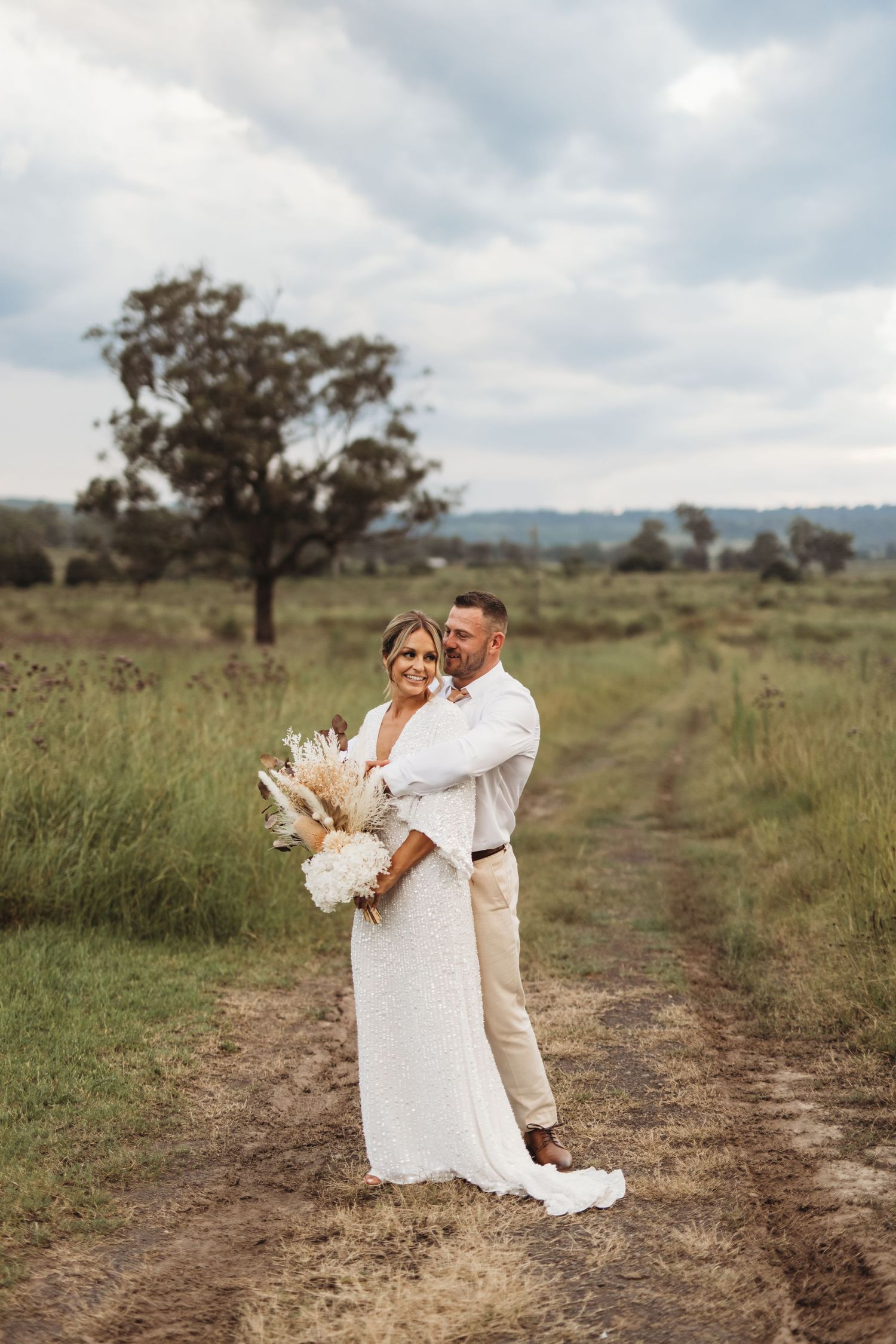 Couple in wedding attire embrace on a dirt path with dried pampas grass bouquet.