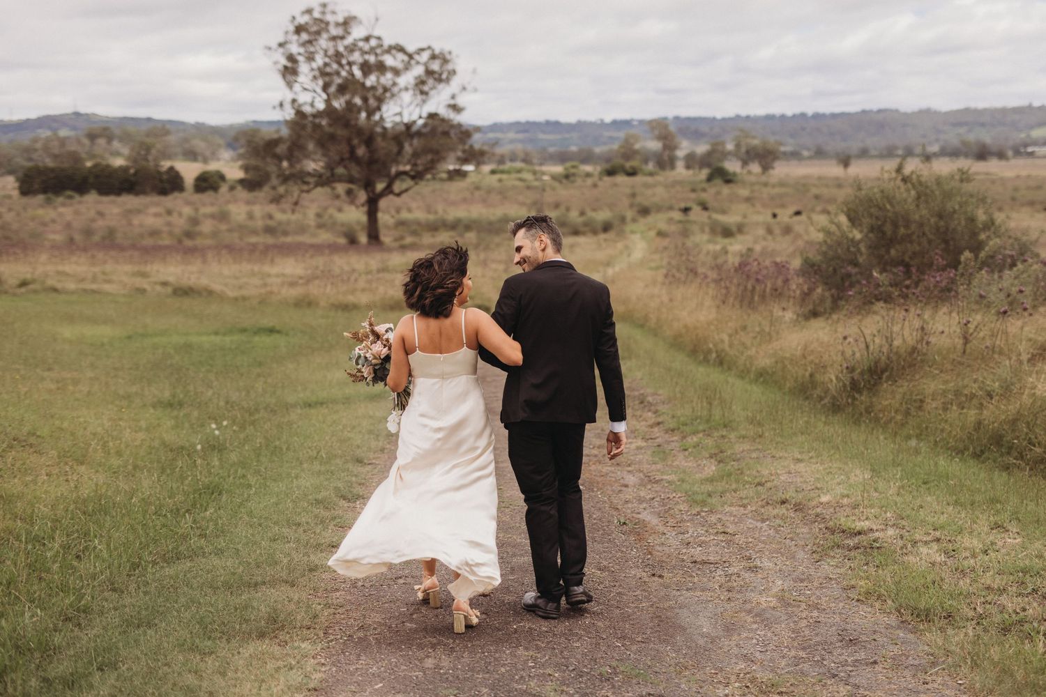 Newlyweds walking away down dirt path through countryside field.