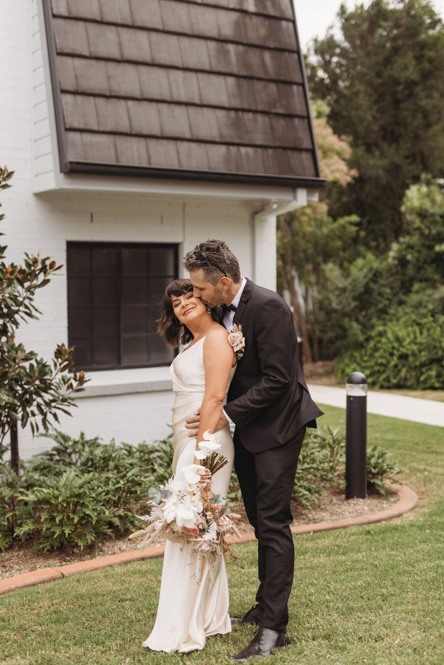 Elegant wedding couple posing together outside white building with landscaped garden.