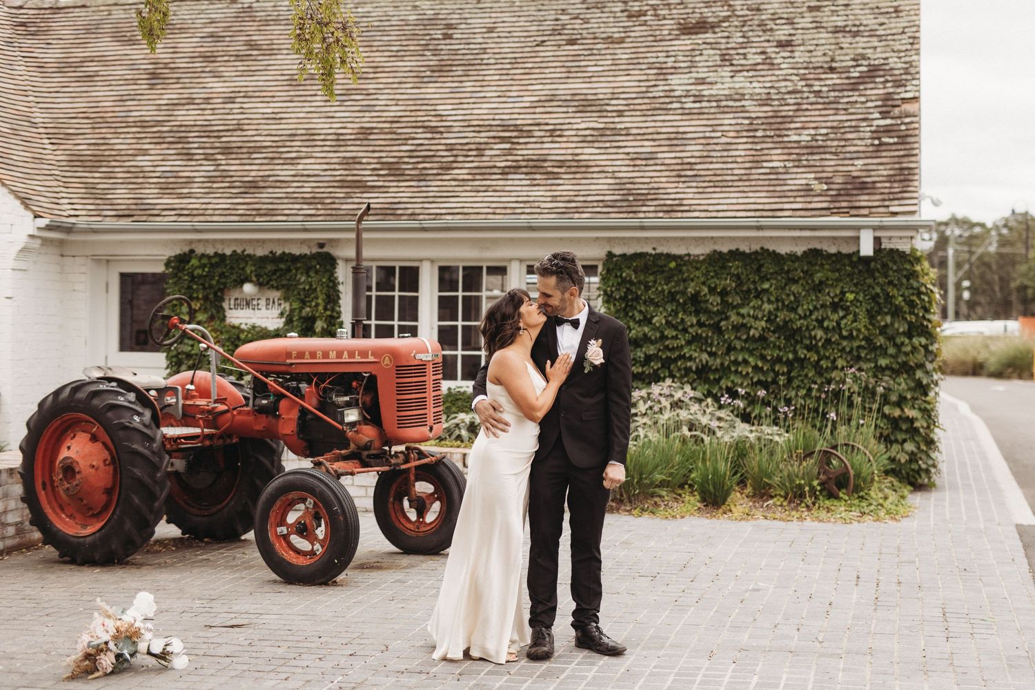 Wedding couple poses by vintage red tractor outside rustic white building.