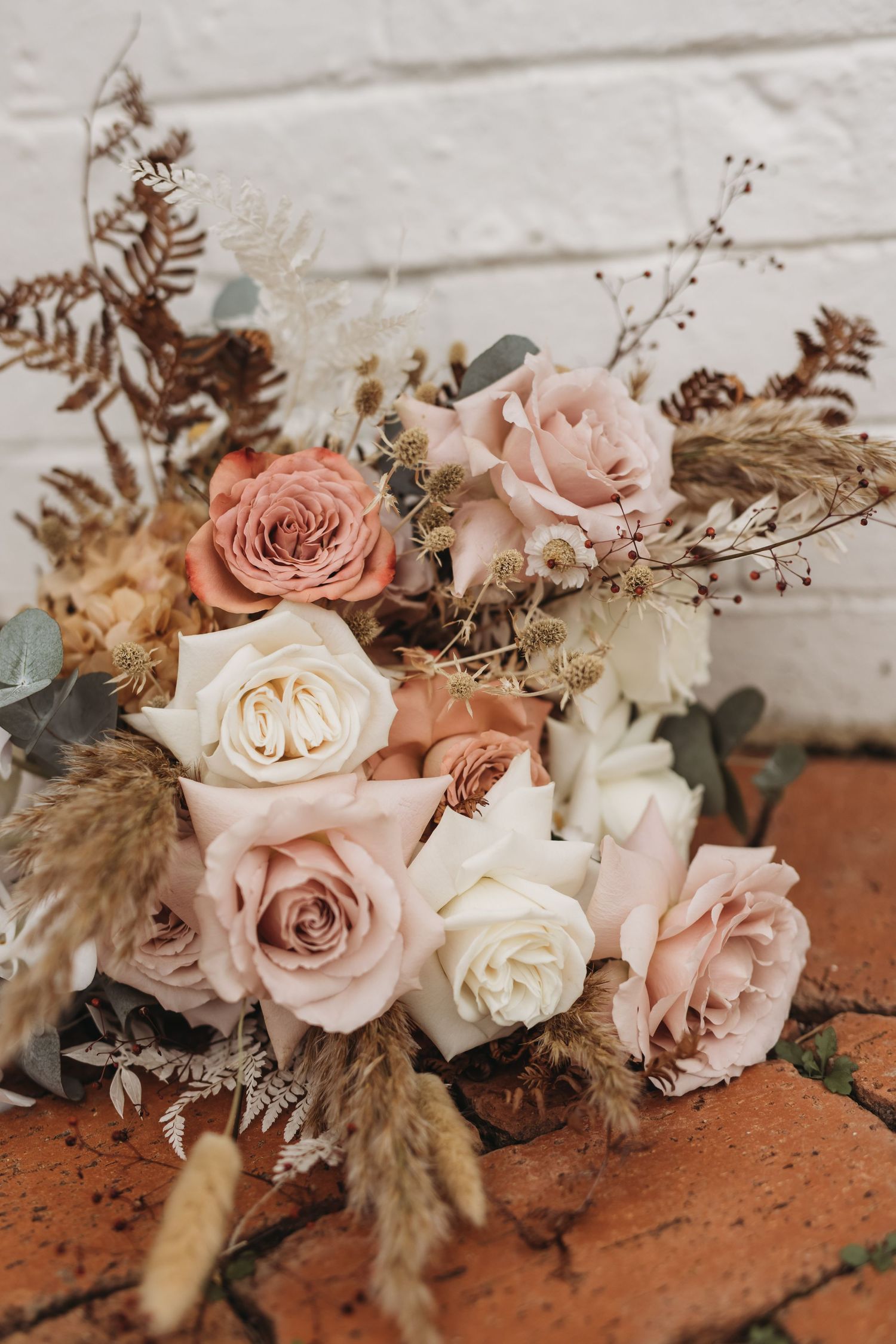 Romantic floral arrangement with blush pink and white roses mixed with dried elements on brick surface.
